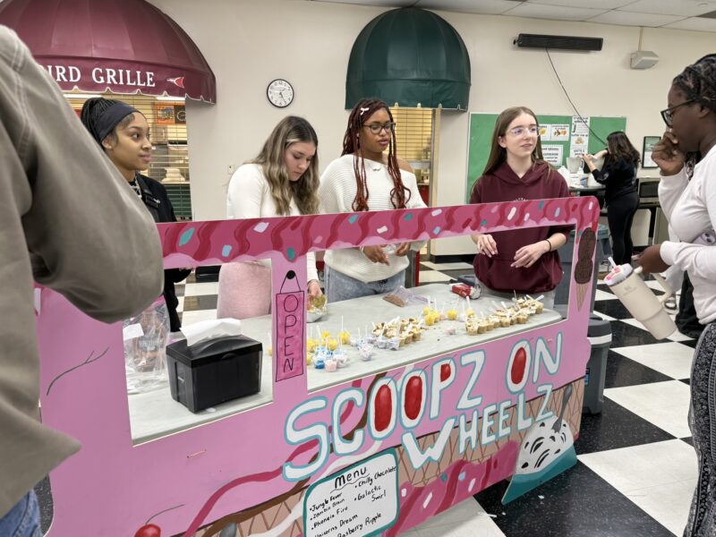 A group of students enjoying work-based learning stands behind a pink ice cream stand marked 